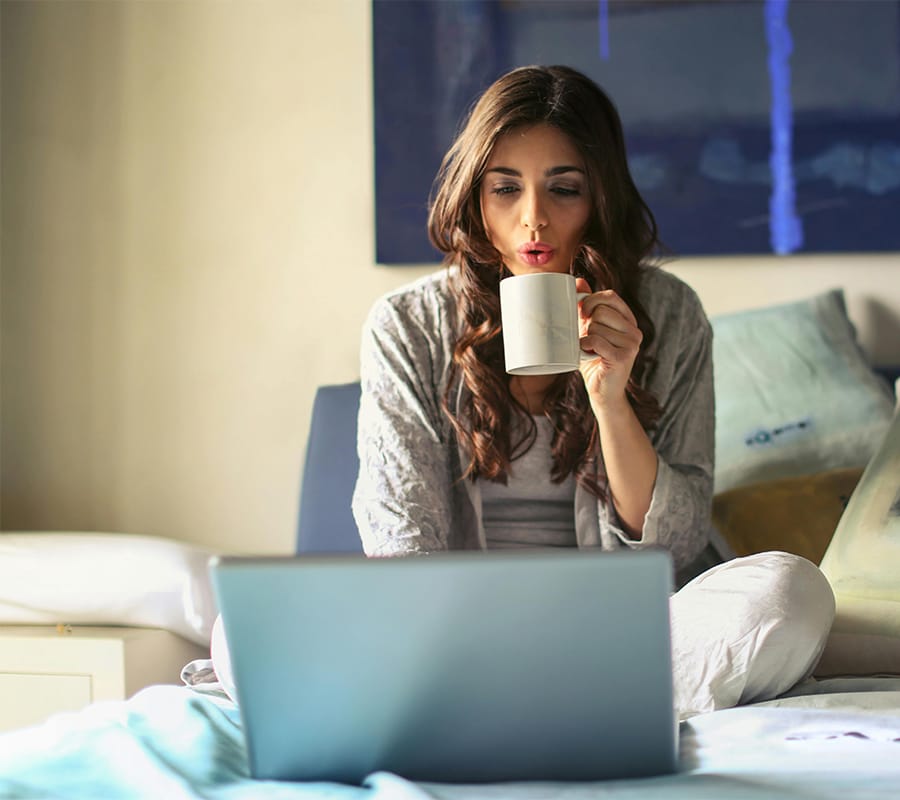 The morning brief Woman sipping her morning coffee on the bed whilst catching up with work on her laptop
